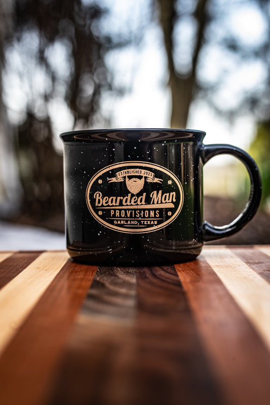 Black mug with 'Bearded Man Provisions' logo on a wooden surface with blurred natural background